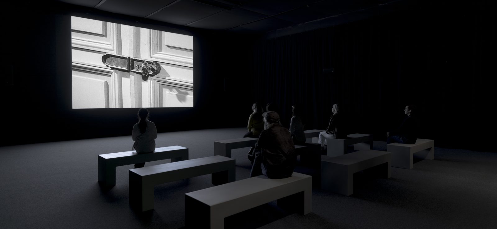People sit on minimalist benches in a dark room, facing a large screen displaying a black-and-white image of a locked door.
