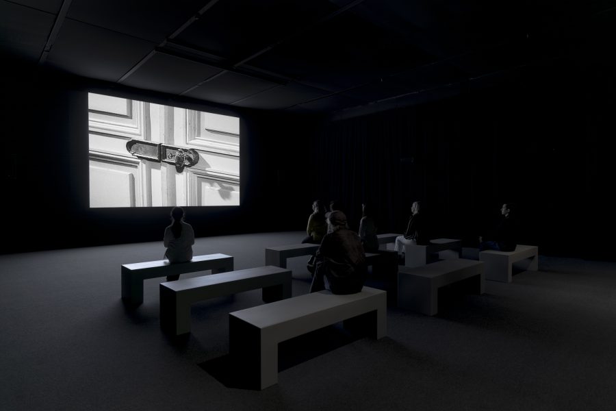 People sit on minimalist benches in a dark room, facing a large screen displaying a black-and-white image of a locked door.