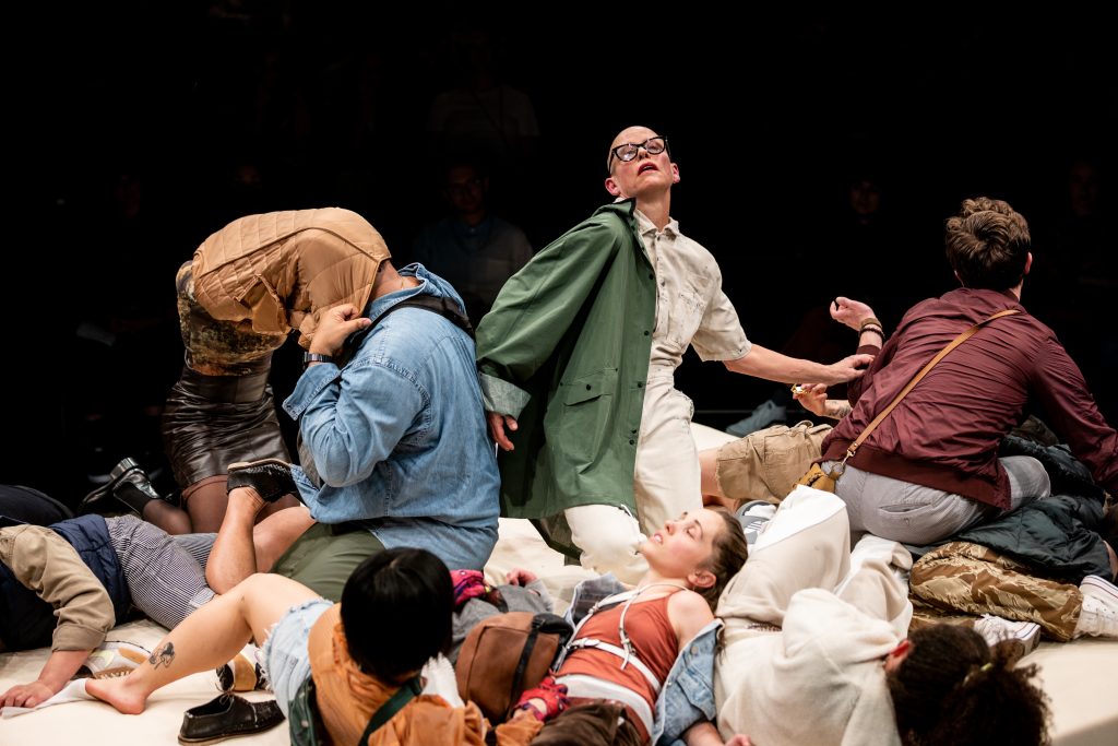 A cluster of people in casual clothing writhe together and on top of each other in a stage setting