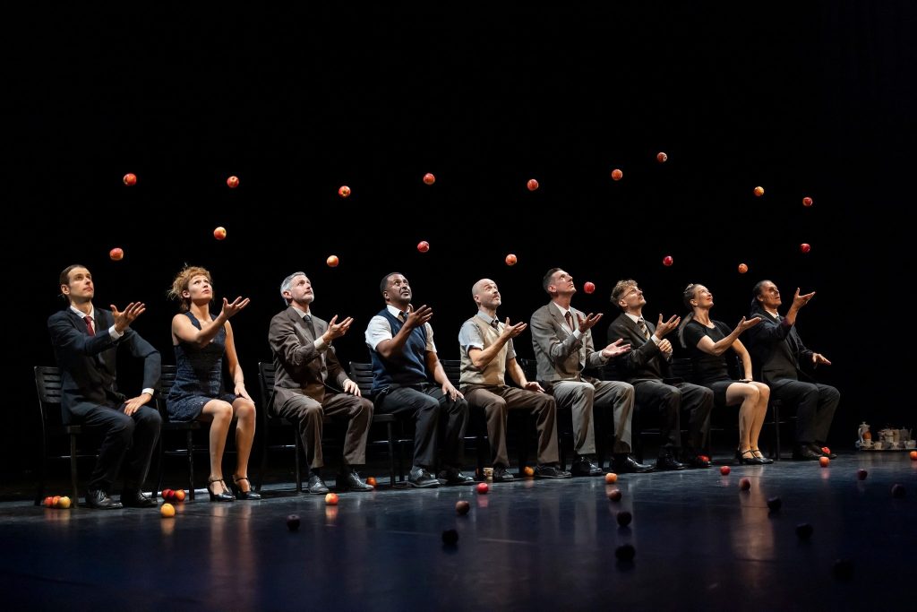 A row of performers sitting on a stage, mid-juggle