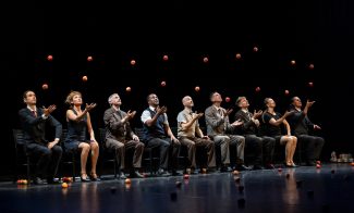 A row of performers sitting on a stage, mid-juggle