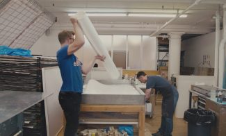 Two people work in a print studio. One person in a blue shirt lifts a large sheet of paper, while the other leans over a printing table. The room is filled with art supplies, and theres a stack of papers on the left.