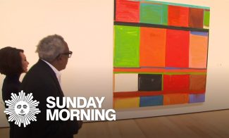 Two people stand in an art gallery looking at a large abstract painting with colorful geometric shapes. The Sunday Morning logo is visible in the corner.