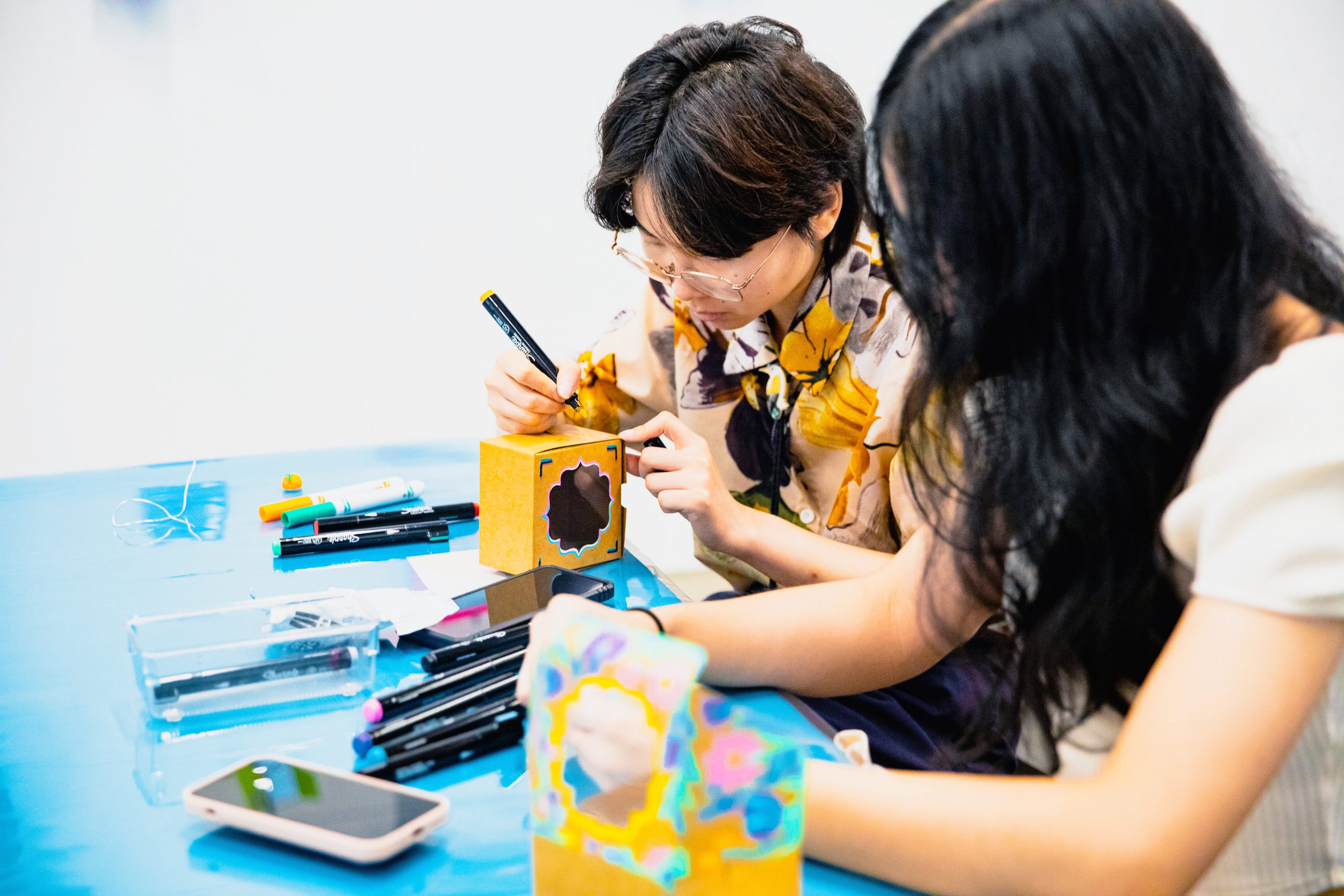 Two medium skinned people sit at a table with a blue table cloth making art using boxes, reflective materials, and markers