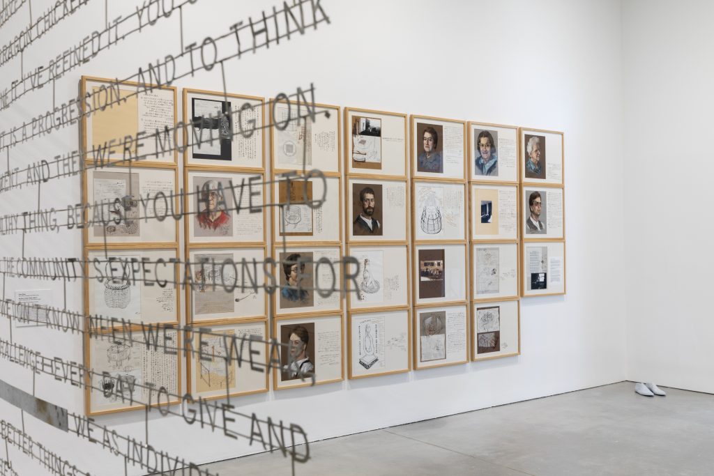 Art exhibition featuring framed sketches, notes, and portraits on a wall. Suspended metal text partially obstructs the view. The floor is smooth concrete, and a pair of cast white shoes is placed in the corner.