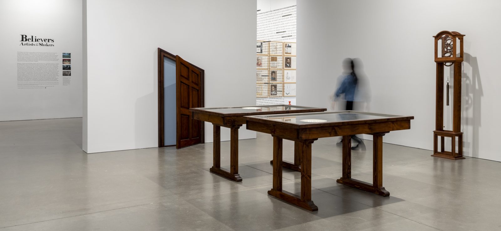 A gallery with 2 wooden tables, a tall wooden clock, and a slanted wooden door embedded in the wall. A blurred figure walks by.