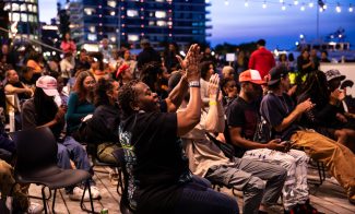 A diverse group of people sit outdoors at dusk, clapping and enjoying a lively Black Joy Day 2024 event under string lights, with city buildings in the background.