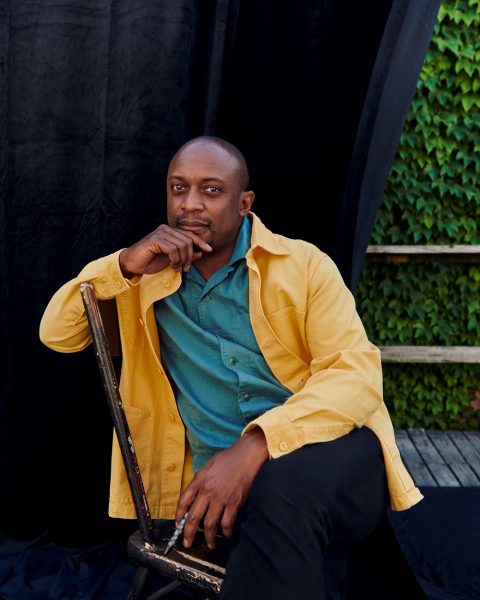 Artist Hank Willis Thomas in bright yellow and blue collared shirts sitting outside on a bench