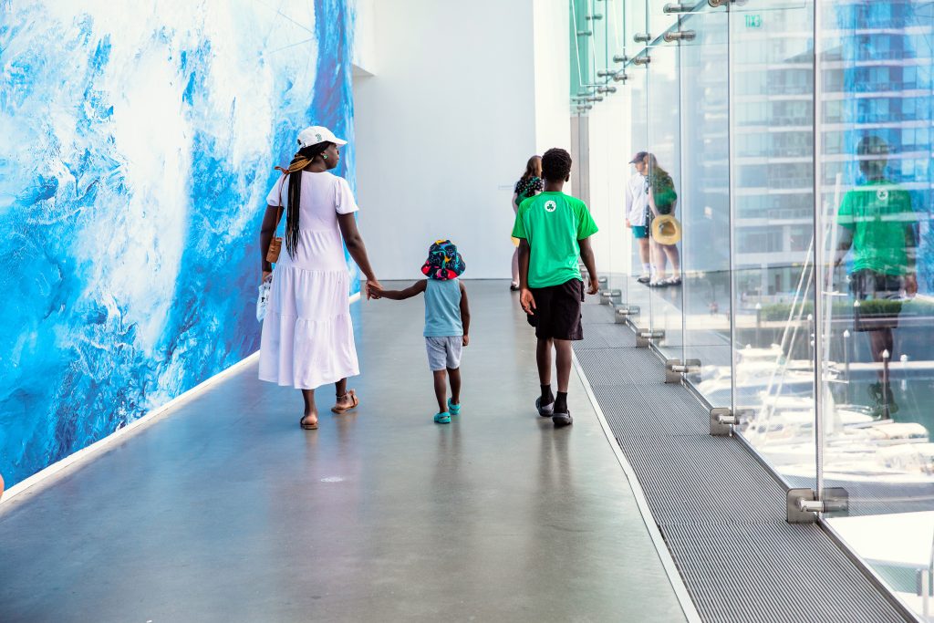 A family of three in casual summer clothes walk down a hallway with large glass windows on one side and a blue mural on the other