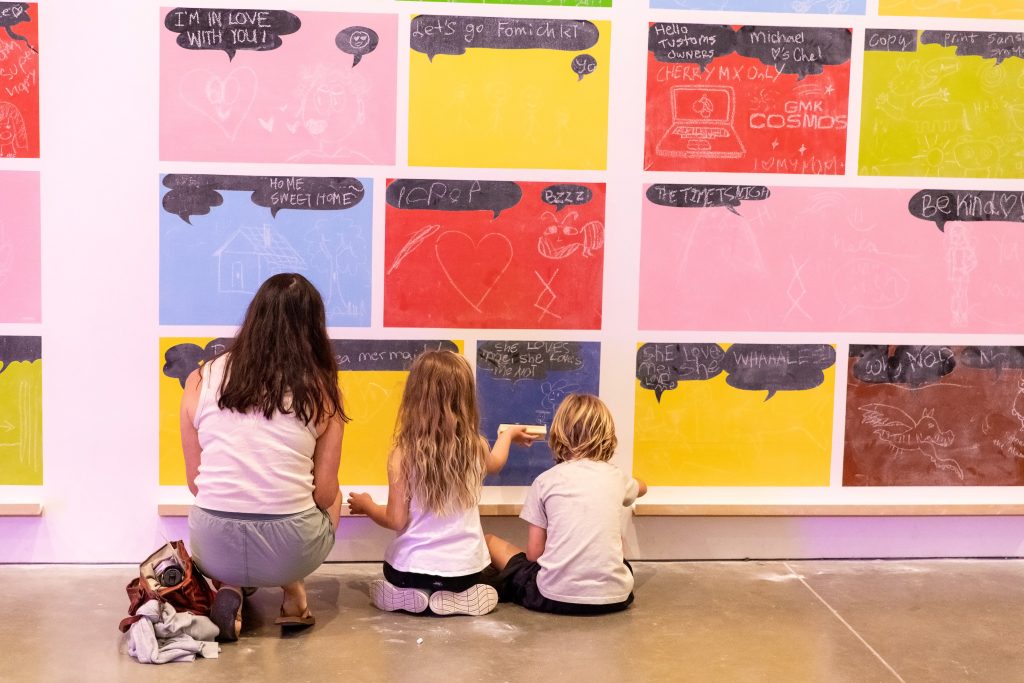 An adult and two children sit on the floor to draw on colorful chalkboard wall tiles