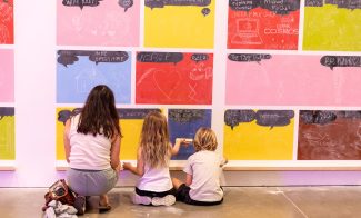 An adult and two children sit on the floor to draw on colorful chalkboard wall tiles