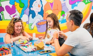 A small child makes art with 2 parents in front of a colorful painted mural