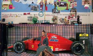 A young adult in a military jacket and skirt sits in front of a red Ferrari race car positioned on a gridded backdrop. Above is a wall-sized collage of found images from magazines.