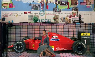 A young adult in a military jacket and skirt sits in front of a red Ferrari race car positioned on a gridded backdrop. Above is a wall-sized collage of found images from magazines.