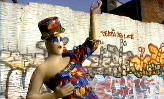 A person in a tan, latex peanut suit wearing a colorful outfit poses in front of a graffiti wall