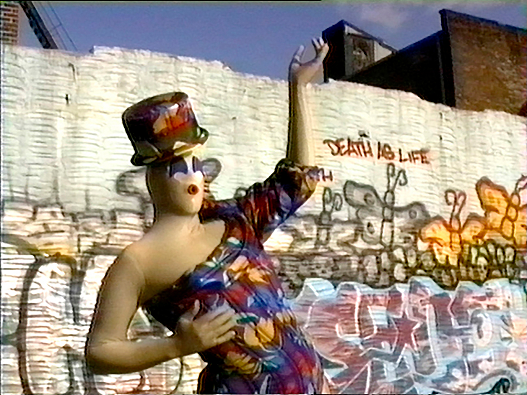 A person in a tan, latex peanut suit wearing a colorful outfit poses in front of a graffiti wall