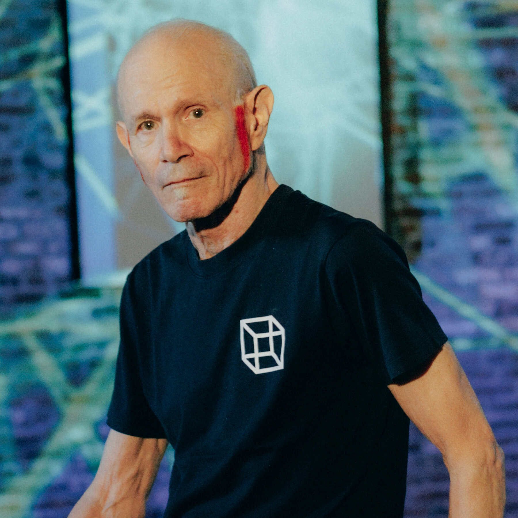 Artist Charles Atlas with orange dyed sideburns and black t-shirt in front of blue background