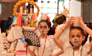 Some children in white dresses with sugar skull face paint pose in front of an altar