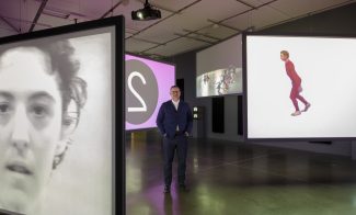 Curator Jeffrey De Blois standing in a dark gallery amidst 4 large video screens playing different footage at the same time