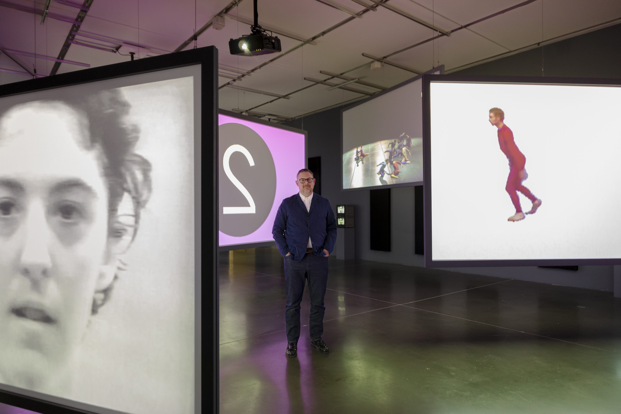 Curator Jeffrey De Blois standing in a dark gallery amidst 4 large video screens playing different footage at the same time