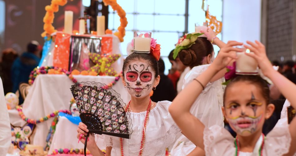 Some children in white dresses with sugar skull face paint pose in front of an altar