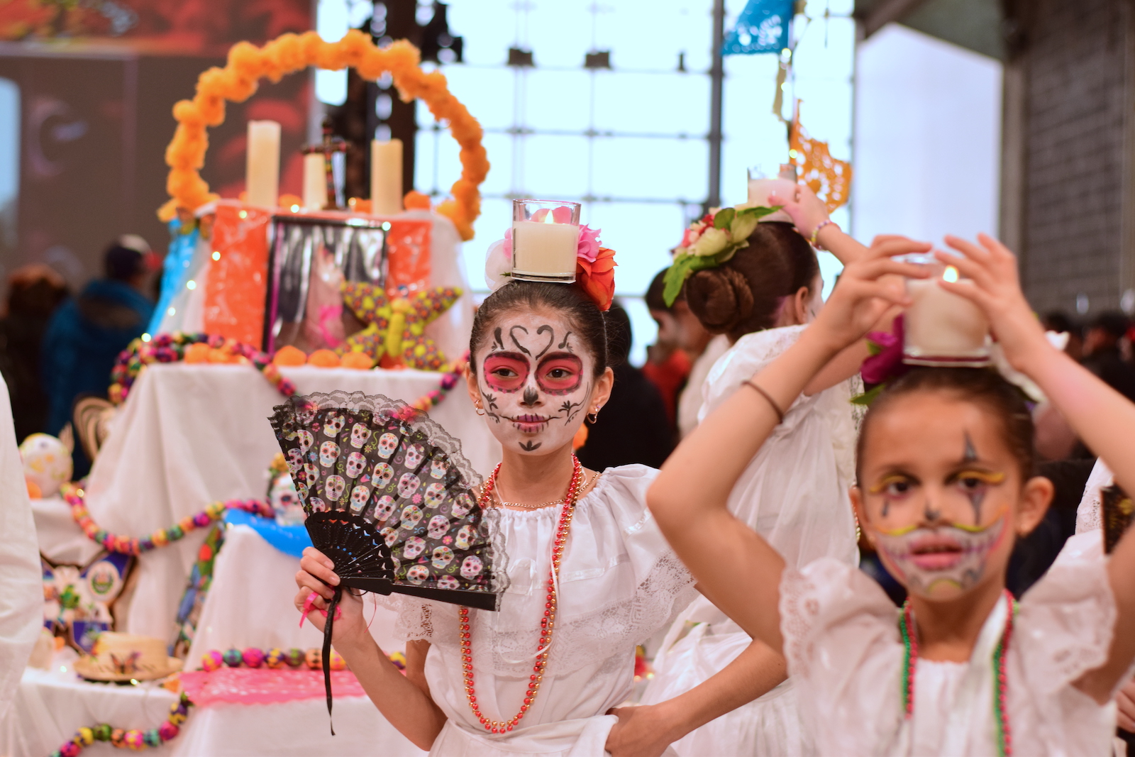 Some children in white dresses with sugar skull face paint pose in front of an altar