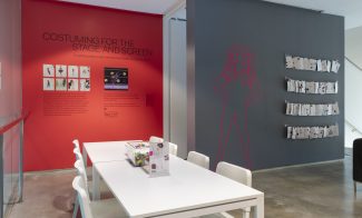 A room with a reddish wall reading "Costuming for the Stage and Screen," a white table, and a gray wall with the lifesize outline of a figure.