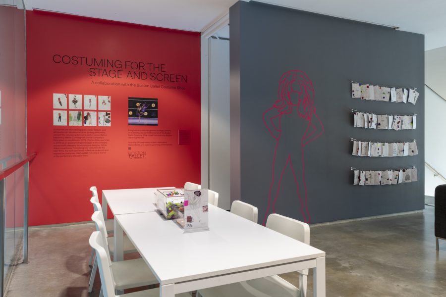 A room with a reddish wall reading "Costuming for the Stage and Screen," a white table, and a gray wall with the lifesize outline of a figure.