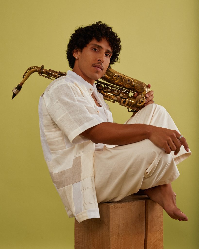 Person in white clothes seated and holding saxophone against yellow background