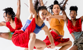 Four dancers leap energetically with expressive movements. They wear colorful outfits, including red and orange costumes. Their expressions are focused and dynamic with hair flying. The background is a plain white, emphasizing their movements.