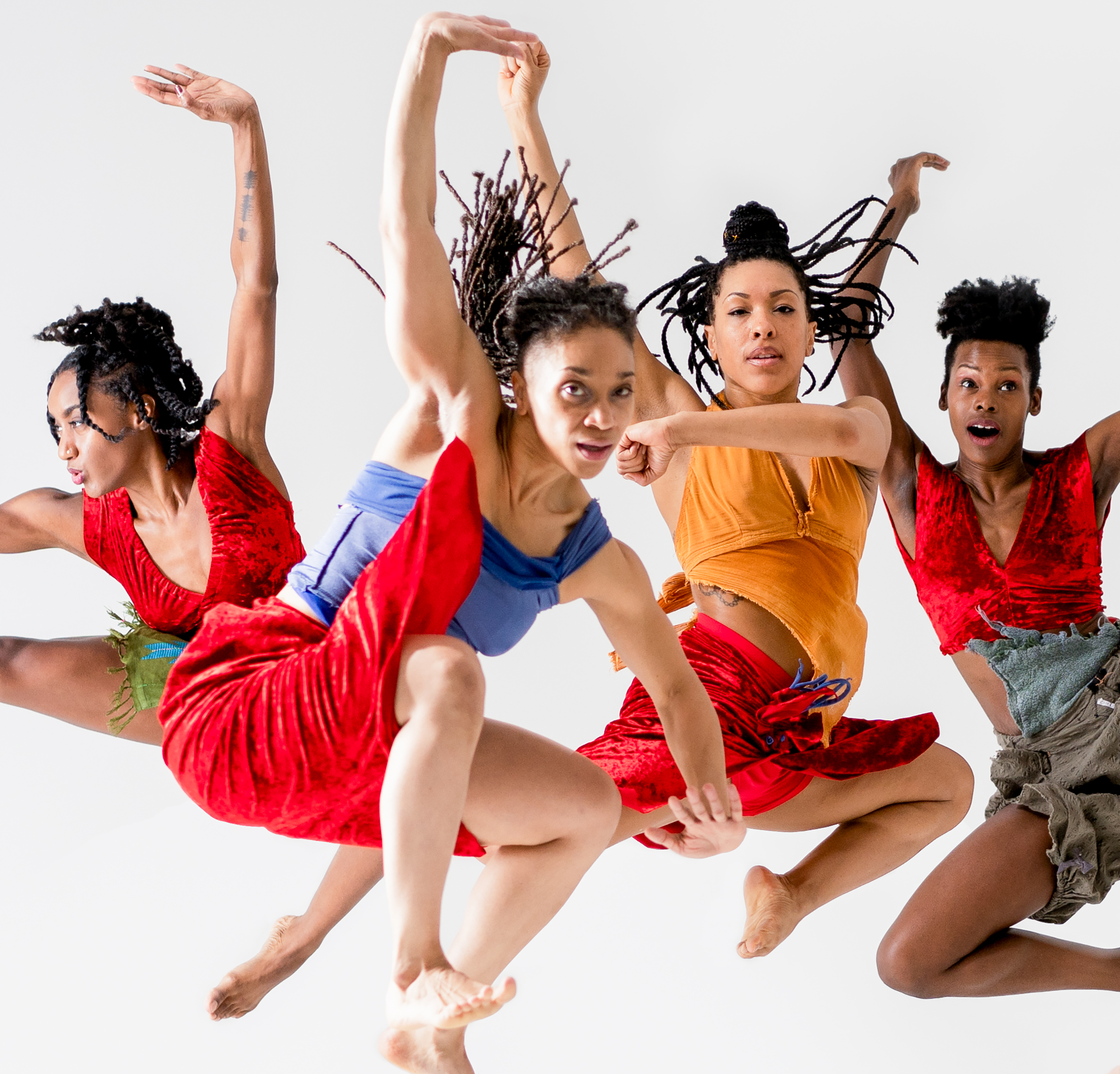 Four dancers leap energetically with expressive movements. They wear colorful outfits, including red and orange costumes. Their expressions are focused and dynamic with hair flying. The background is a plain white, emphasizing their movements.