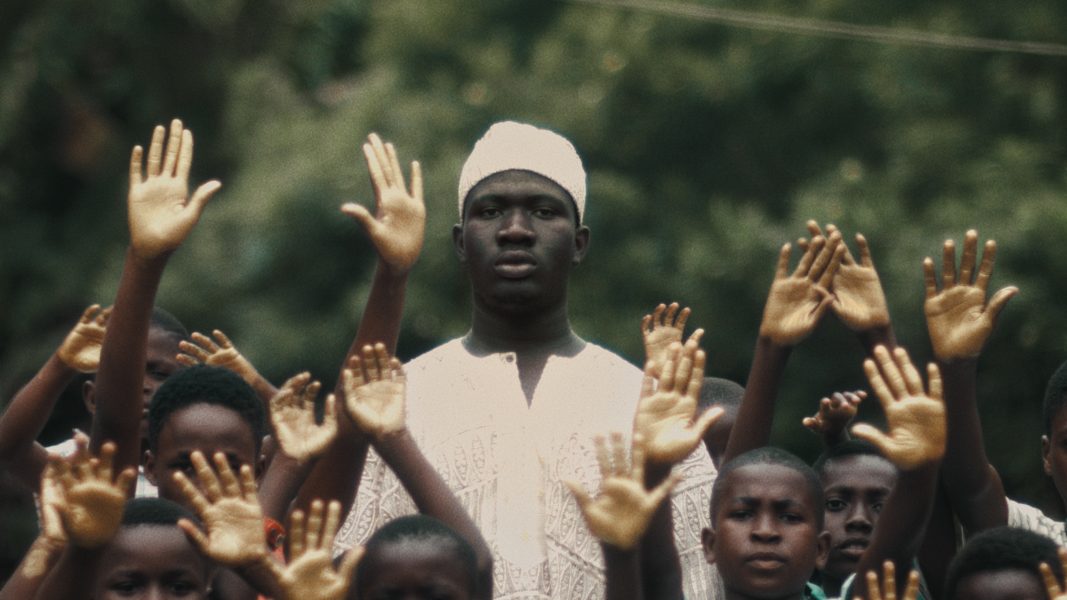 A man in traditional white clothing and a hat stands among a crowd, their hands raised, set against lush green foliage.