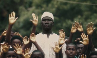 A man in traditional white clothing and a hat stands among a crowd, their hands raised, set against lush green foliage.