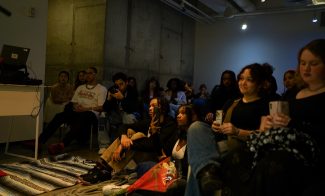 A diverse group of people sit in a dimly lit room, attentively watching something out of view. Some hold phones, and a few sit on the floor. The atmosphere appears relaxed and focused.