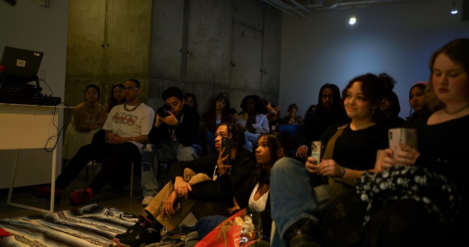 A diverse group of people sit in a dimly lit room, attentively watching something out of view. Some hold phones, and a few sit on the floor. The atmosphere appears relaxed and focused.