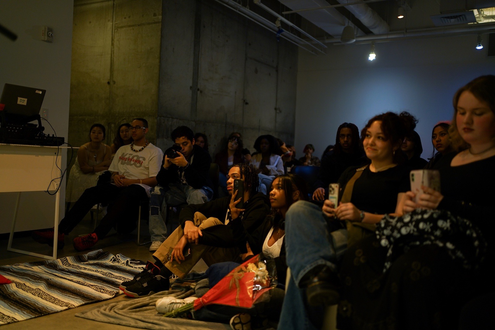 A diverse group of people sit in a dimly lit room, attentively watching something out of view. Some hold phones, and a few sit on the floor. The atmosphere appears relaxed and focused.