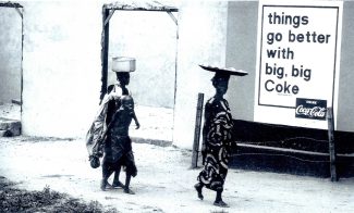 Two people walk by a wall with a Coca-Cola advertisement that reads, things go better with big, big Coke. One carries a bowl on their head, the other carries a bundle on their back.