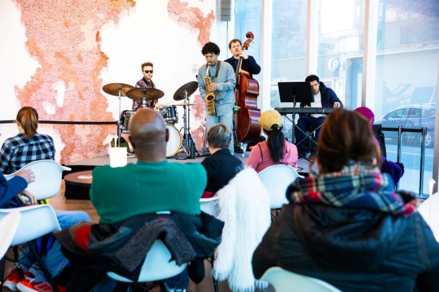 A jazz quartet, reminiscent of the Gregory Groover Jr Trio, performs in a modern venue. The musicians play drums, saxophone, double bass, and keyboard. An audience in casual attire listens intently as light streams through large windows onto coral-colored wall art.