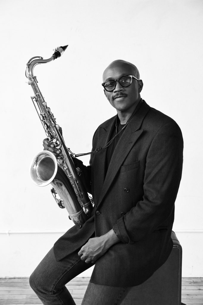 A person in a suit sits while holding a saxophone. They are wearing glasses and have a calm expression. The background is a plain, light-colored wall, creating a minimalist setting.