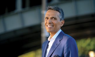 A person in a blue suit smiles while standing outdoors. A blurred structure is visible in the background.