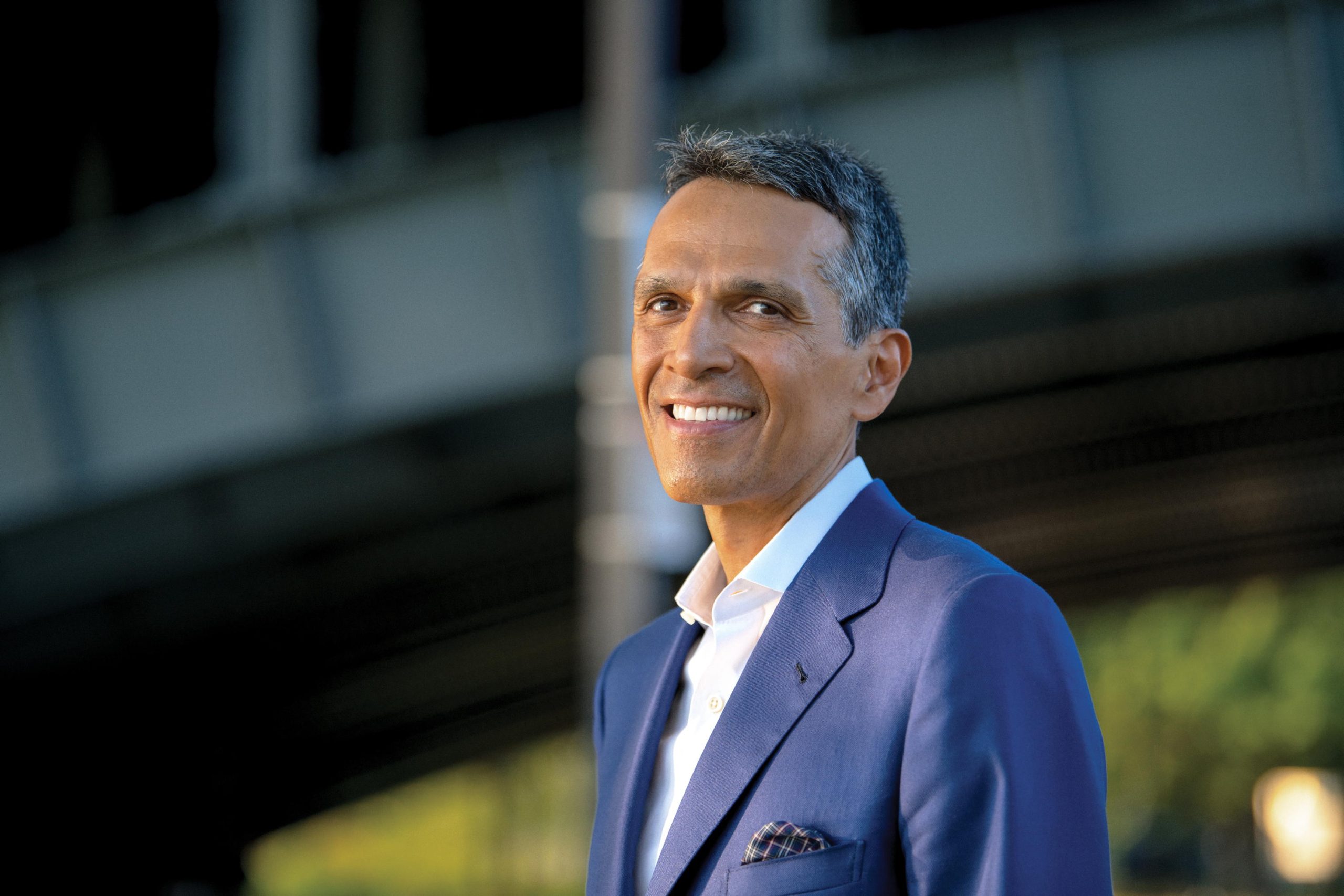 A person in a blue suit smiles while standing outdoors. A blurred structure is visible in the background.