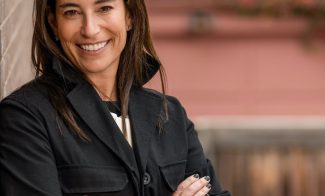 A woman with long dark hair is smiling and standing with her arms crossed, wearing a black jacket. She is leaning against a brick wall outdoors, with a blurred background of a building and some greenery.