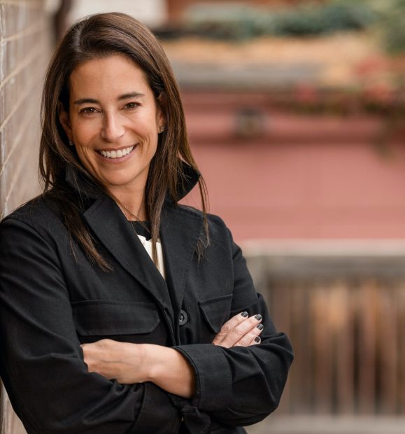 A woman with long dark hair is smiling and standing with her arms crossed, wearing a black jacket. She is leaning against a brick wall outdoors, with a blurred background of a building and some greenery.