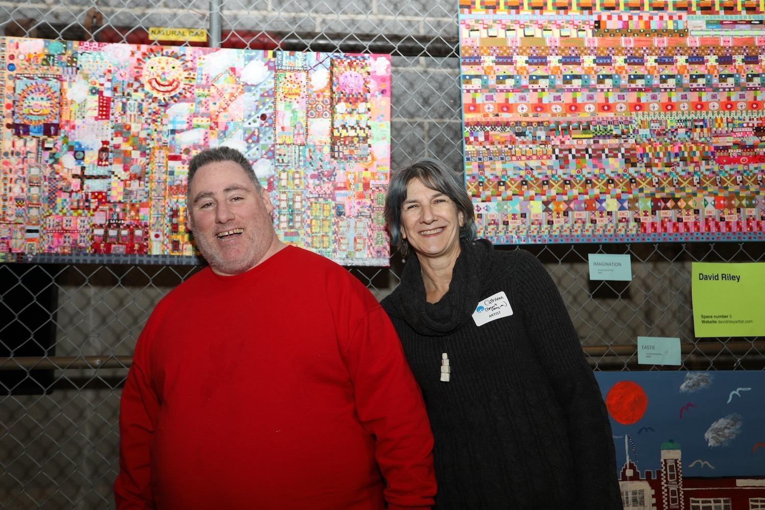 A man in a red sweater and a woman smiling stand in front of colorful abstract artworks displayed on a chain-link fence.