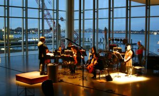 A conductor leads a small orchestra inside a glass-walled building at dusk, overlooking a harbor. The warm lighting highlights the performers against the evening scenery, as musicians play string and wind instruments with sheet music on stands.