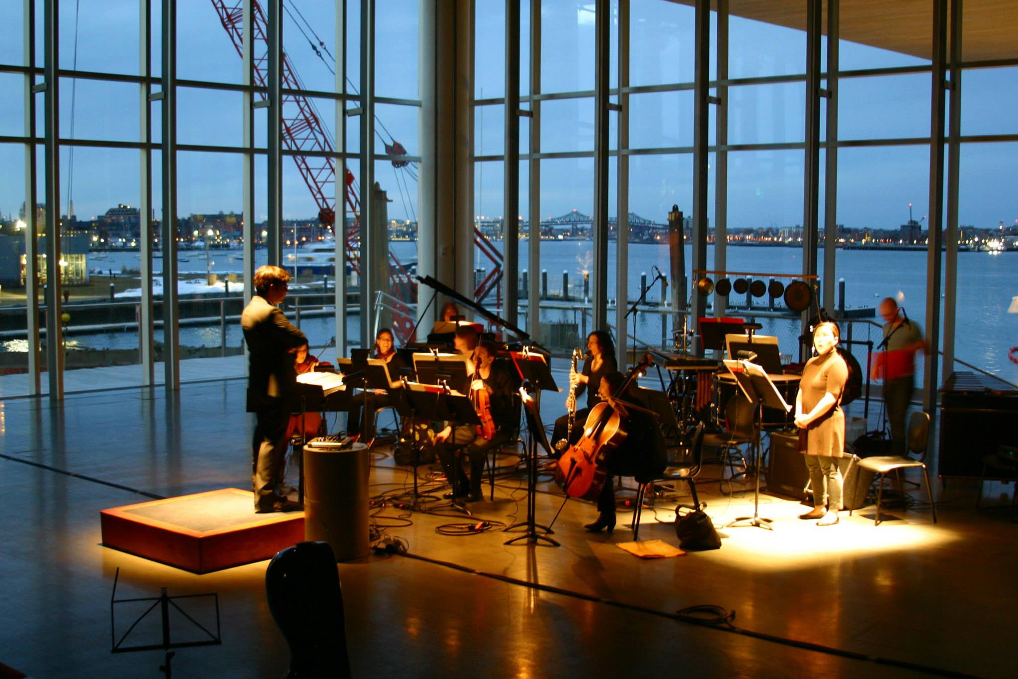 A conductor leads a small orchestra inside a glass-walled building at dusk, overlooking a harbor. The warm lighting highlights the performers against the evening scenery, as musicians play string and wind instruments with sheet music on stands.