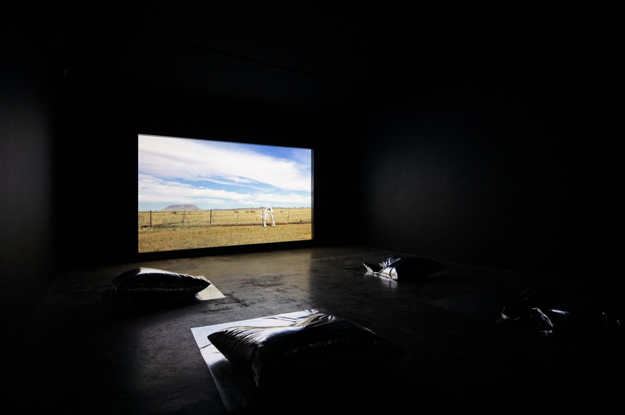 In a dark room, "Star Scores" by Roberto Carlos Lange and Kristi Sword plays softly as a large screen displays a scenic countryside image featuring a horse in a field. Reflective pillows are placed on the floor, inviting viewers to sit and immerse themselves in the tranquil experience.