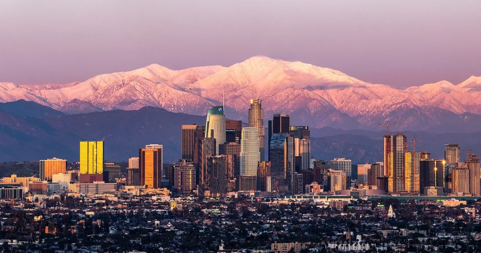 A panoramic view of a city skyline at sunset, with tall skyscrapers glowing in golden light. In the background, snow-capped mountains are illuminated by a soft pink hue, under a clear sky.