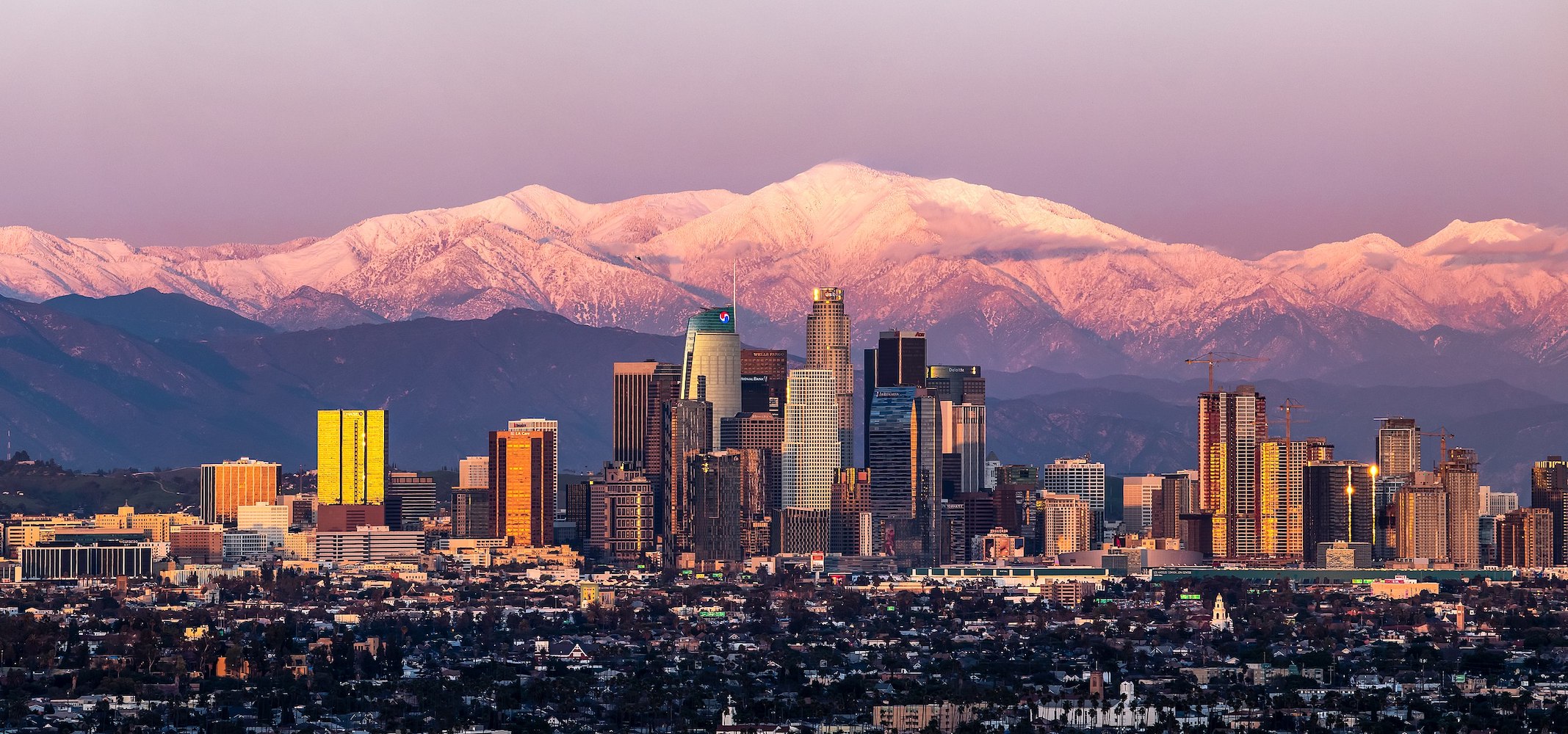 A panoramic view of a city skyline at sunset, with tall skyscrapers glowing in golden light. In the background, snow-capped mountains are illuminated by a soft pink hue, under a clear sky.