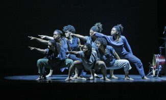 Six dancers perform on stage in a dramatic pose. Clad in casual clothing, they point forward in unison with bent knees, creating a dynamic and coordinated movement against the dark background that highlights their expressive form.